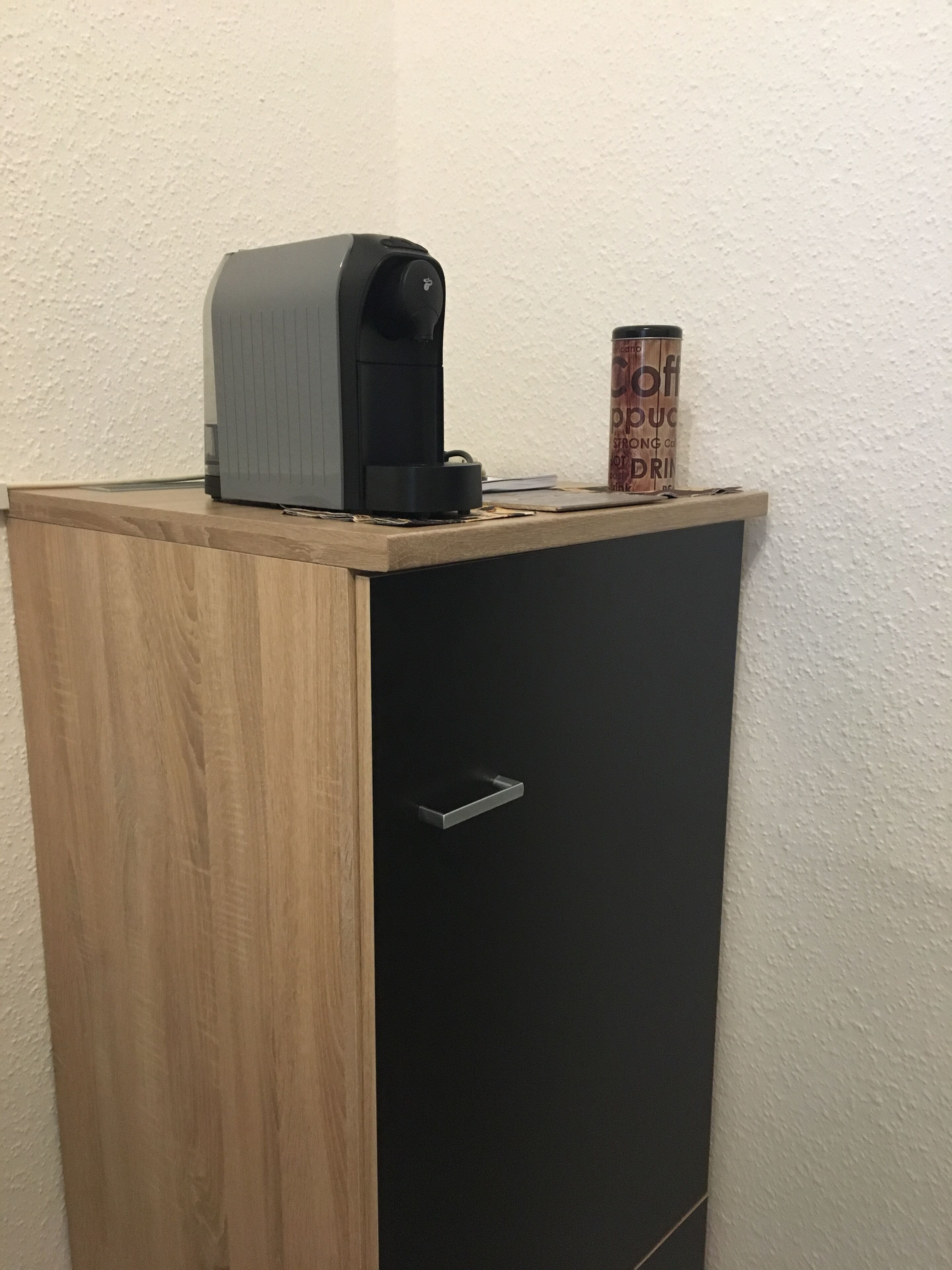 A small wooden cabinet with a black door, a coffee maker on top, and a can of coffee on the side.