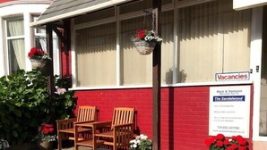 A red and beige building with a 'Vacancies' sign, two wooden chairs, and potted plants. - The Sandalwood (Blackpool)
