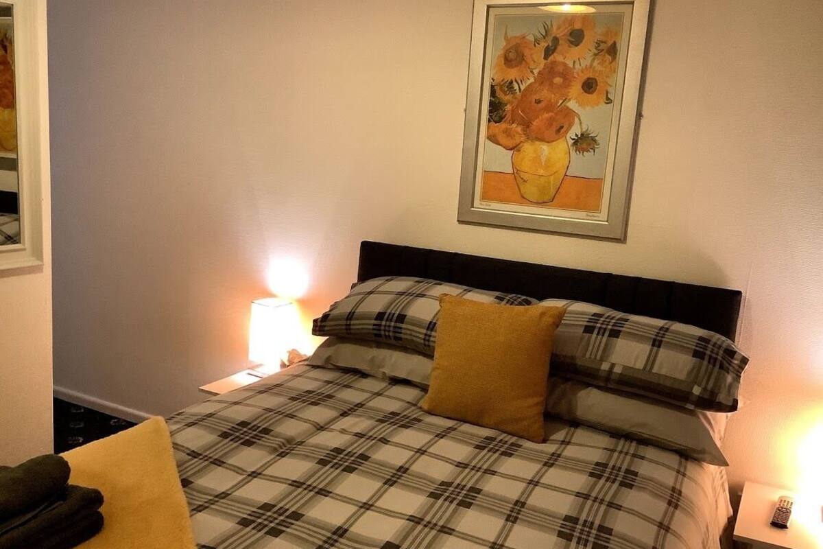 Un lit bien fait, avec une couette à carreaux, un tableau encadré représentant des tournesols accroché au mur et une lampe de chevet.