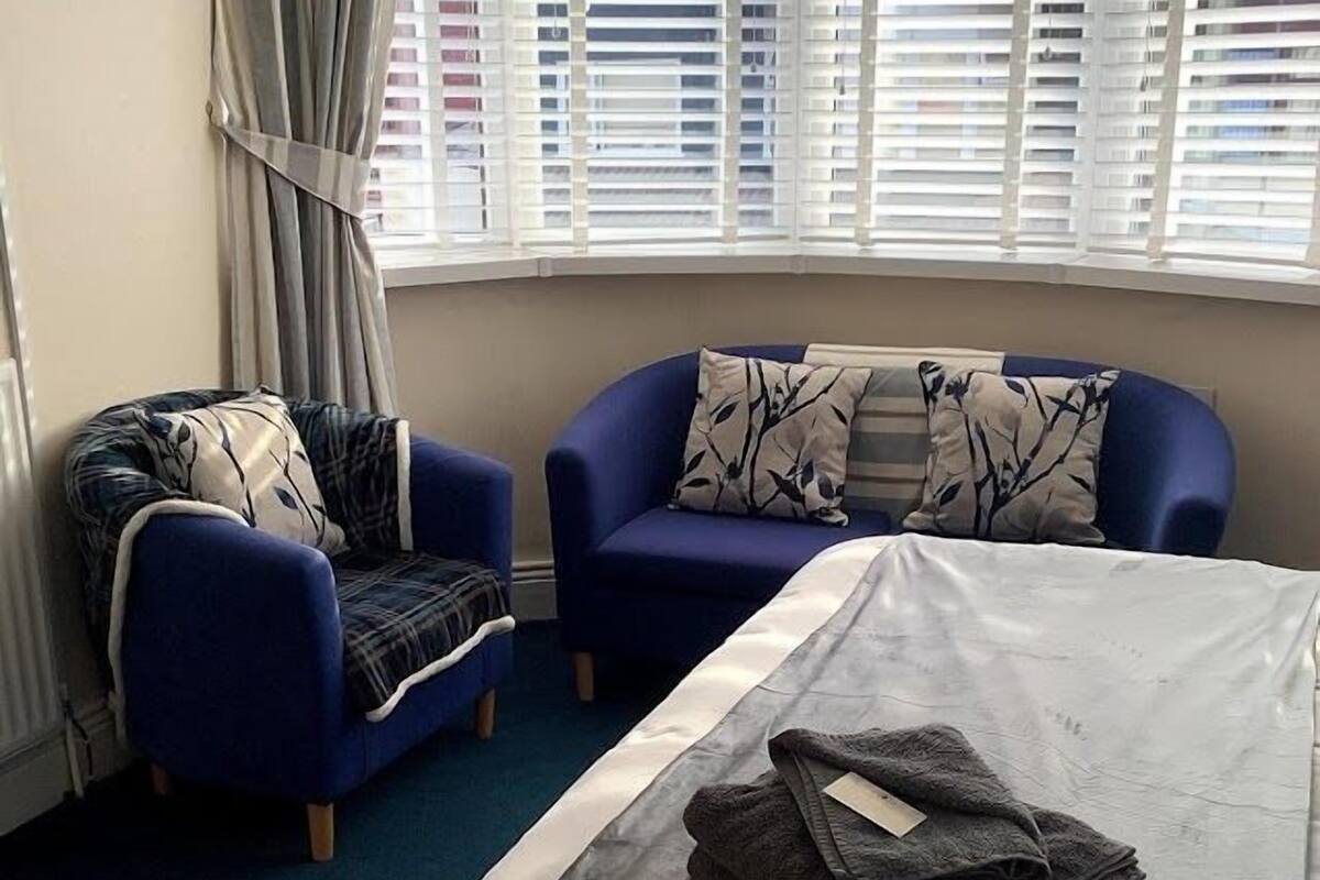 Une chambre d’hôtel avec un lit, deux fauteuils, une fenêtre avec des stores et un rideau.
