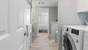 Un lavadero con lavadora y secadora, un mueble pequeño y una puerta que da a un dormitorio.