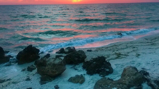 Una playa con rocas en primer plano y un atardecer sobre el océano.