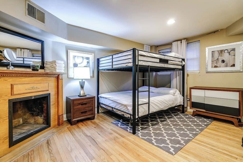 Un dormitorio con una cama litera, un aparador de madera, un espejo, una chimenea y una alfombra estampada.