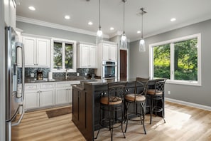 A modern kitchen with white cabinets, a central island, bar stools, and a large window.