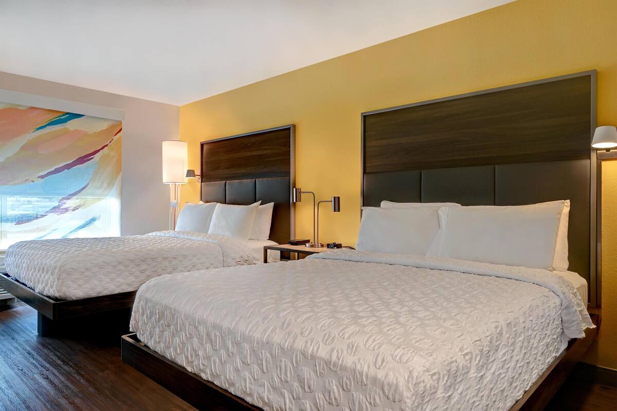Une chambre d’hôtel avec deux lits, chacun doté d’une tête de lit sombre et de draps blancs. On y trouve des tables de chevet avec des lampes, ainsi qu’un grand tableau abstrait accroché au mur.