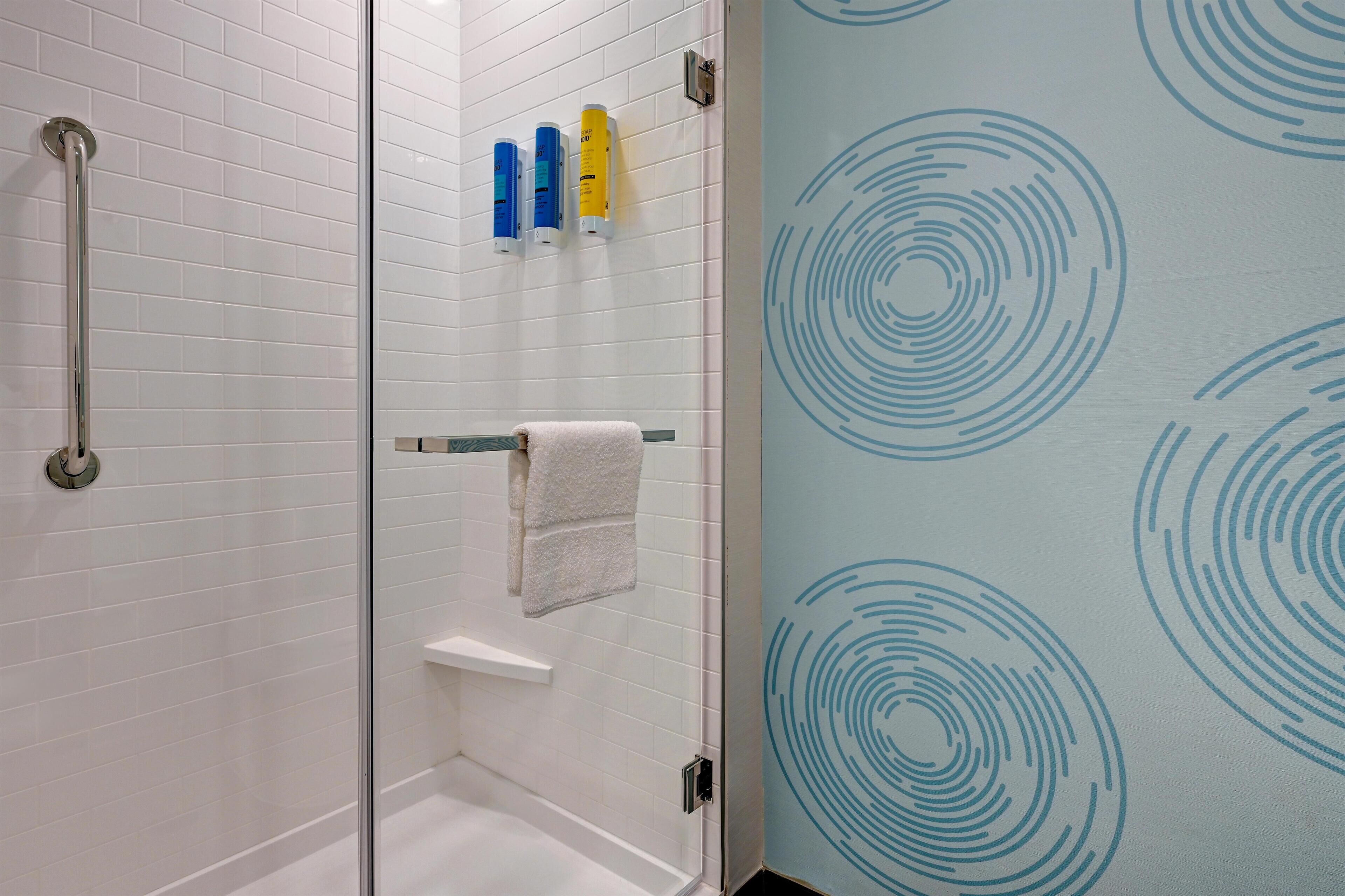 A bathroom with white tiled walls, a glass shower enclosure, a white towel on a rack, and blue circular patterns on the wall.