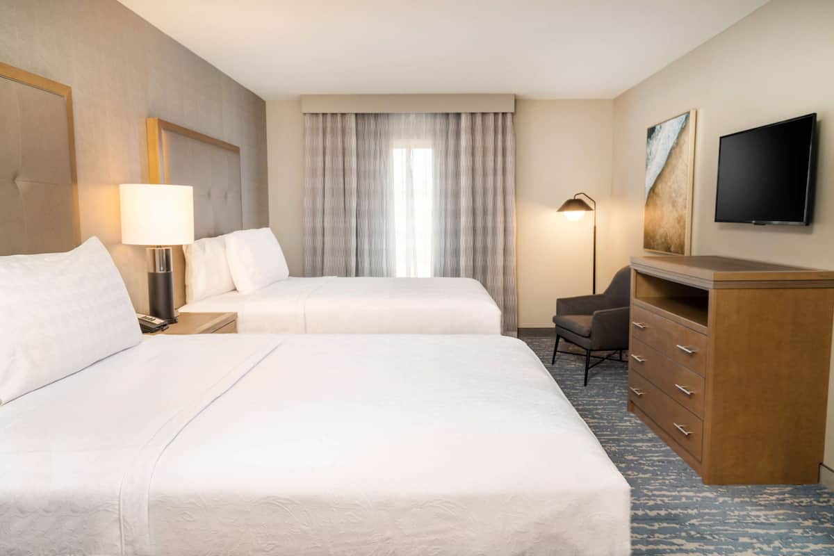 A hotel room with two beds, a desk, a chair, a dresser, a lamp, a TV, and a window with curtains.
