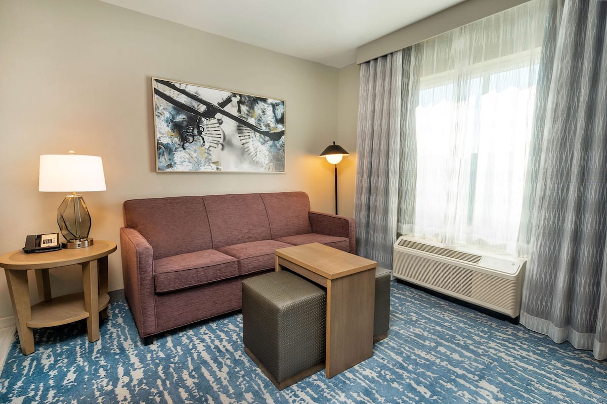 A hotel room with a red sofa, a small wooden table with a lamp, a large abstract painting, and a window with curtains.