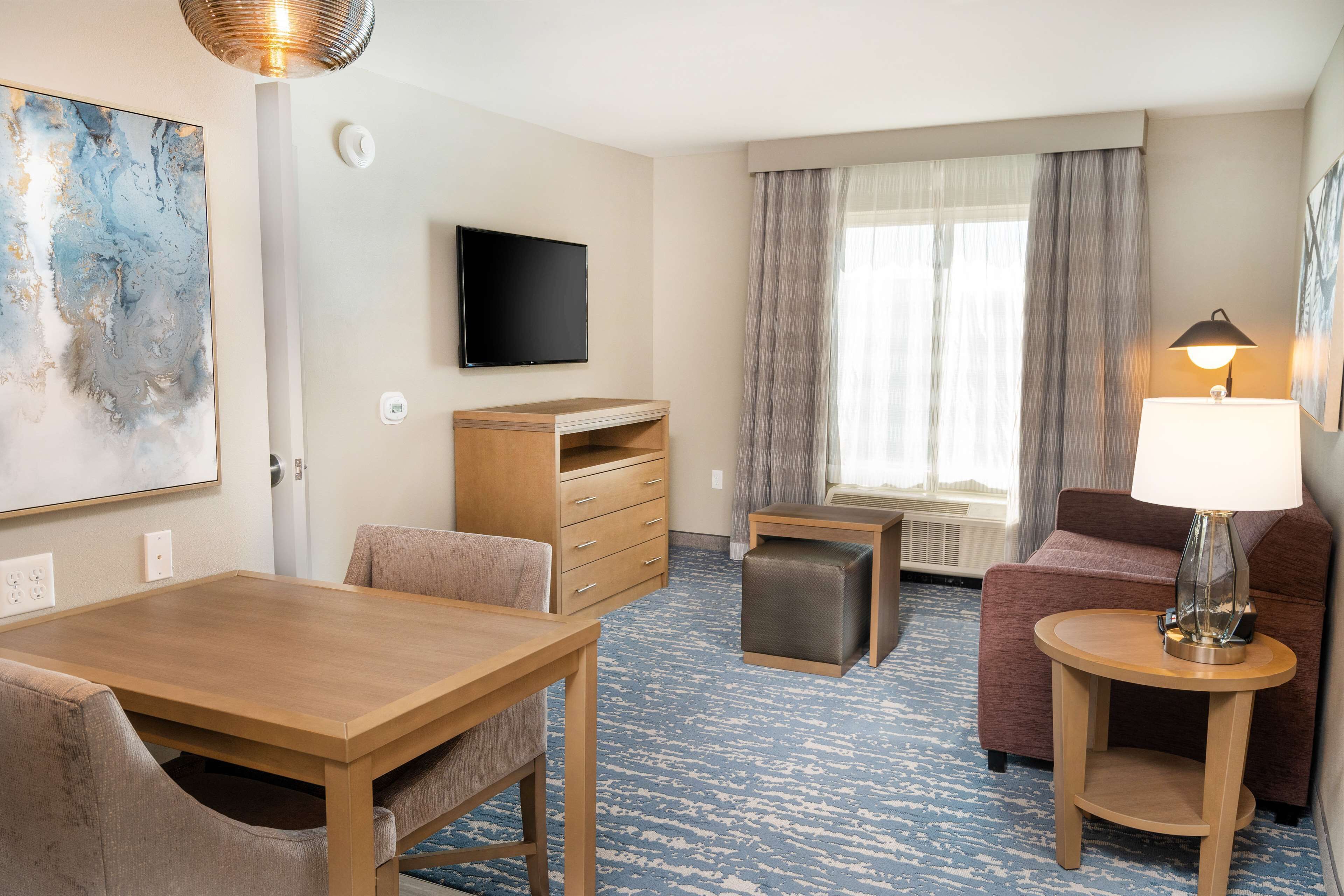 Habitación de hotel con una mesa de comedor de madera, un sofá, un televisor, una lámpara y una ventana con cortinas.