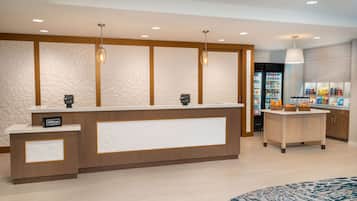 Un espace d’accueil d’hôtel au design moderne, comprenant un comptoir de réception, un minibar et un réfrigérateur rempli de collations.
