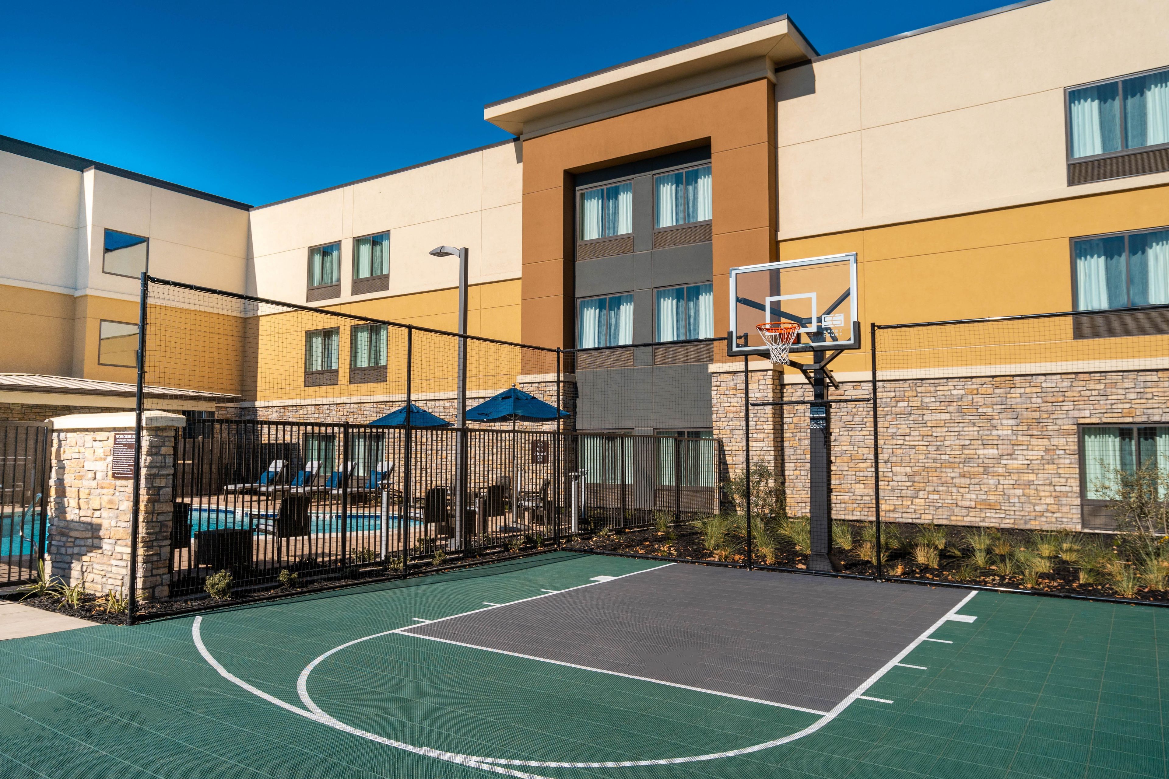 An outdoor basketball court adjacent to a building with a swimming pool and lounge chairs.