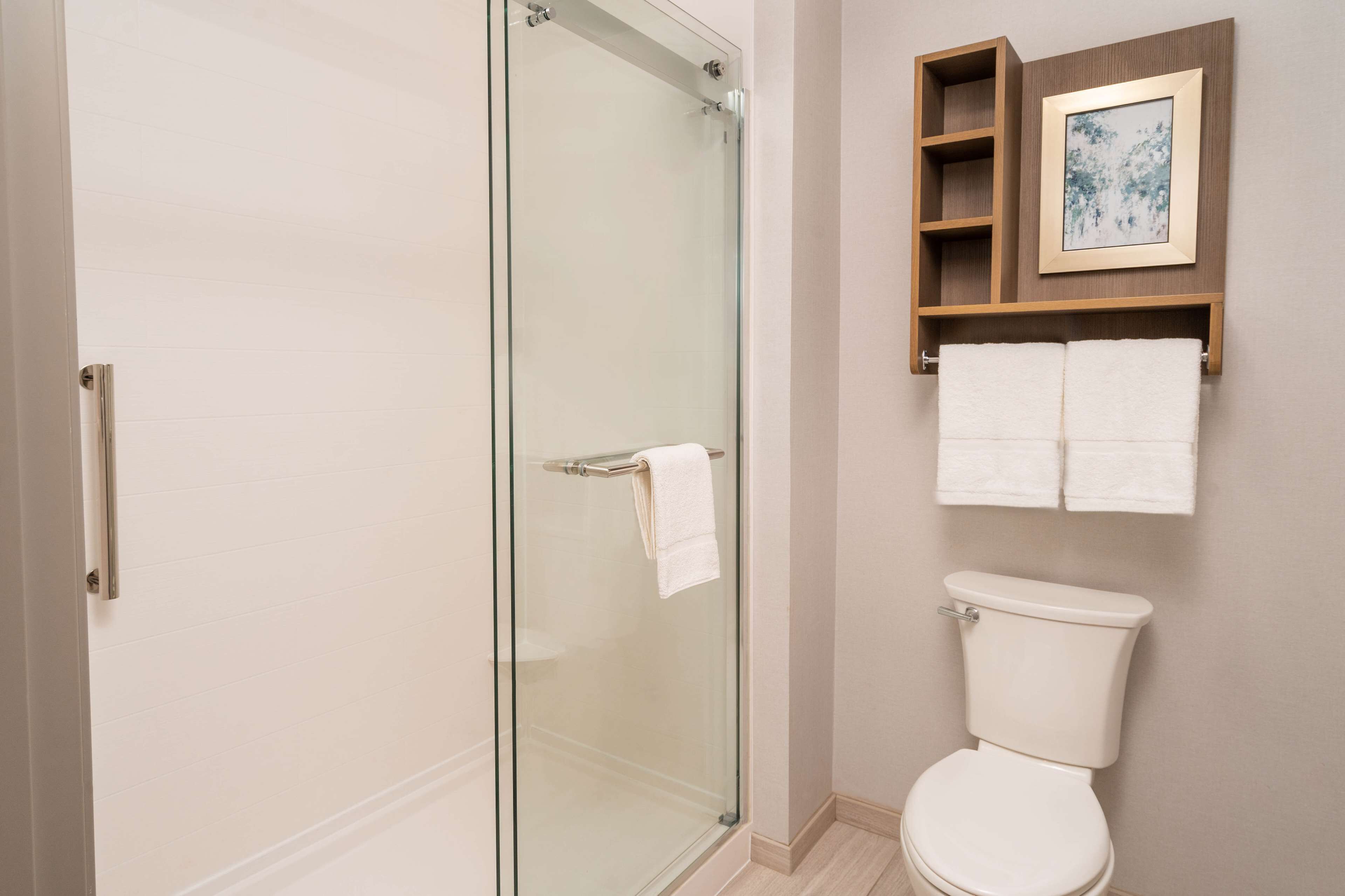 Un baño con cabina de ducha de vidrio, inodoro y repisa con toallas y un cuadro enmarco.