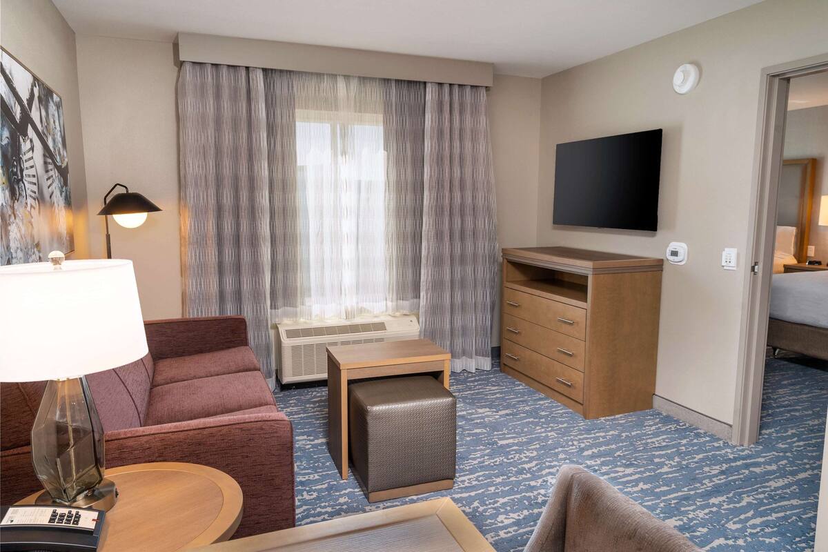 A hotel room with a sofa, a small table with a lamp, a TV mounted on the wall, a wooden dresser, and a bed.