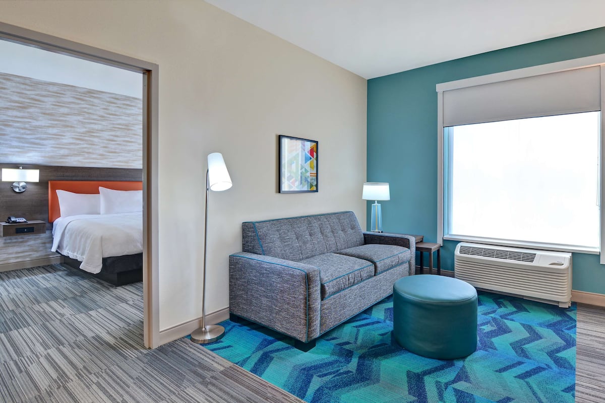 Une chambre d’hôtel avec un lit, un canapé, une petite table, une lampe, un tableau au mur et une grande fenêtre.