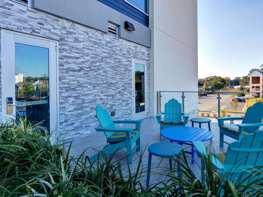 Un edificio moderno con fachada de piedra, zona de estar al aire libre con sillas y mesas tipo Adirondack azules, y vista a un estacionamiento y otros edificios al fondo.