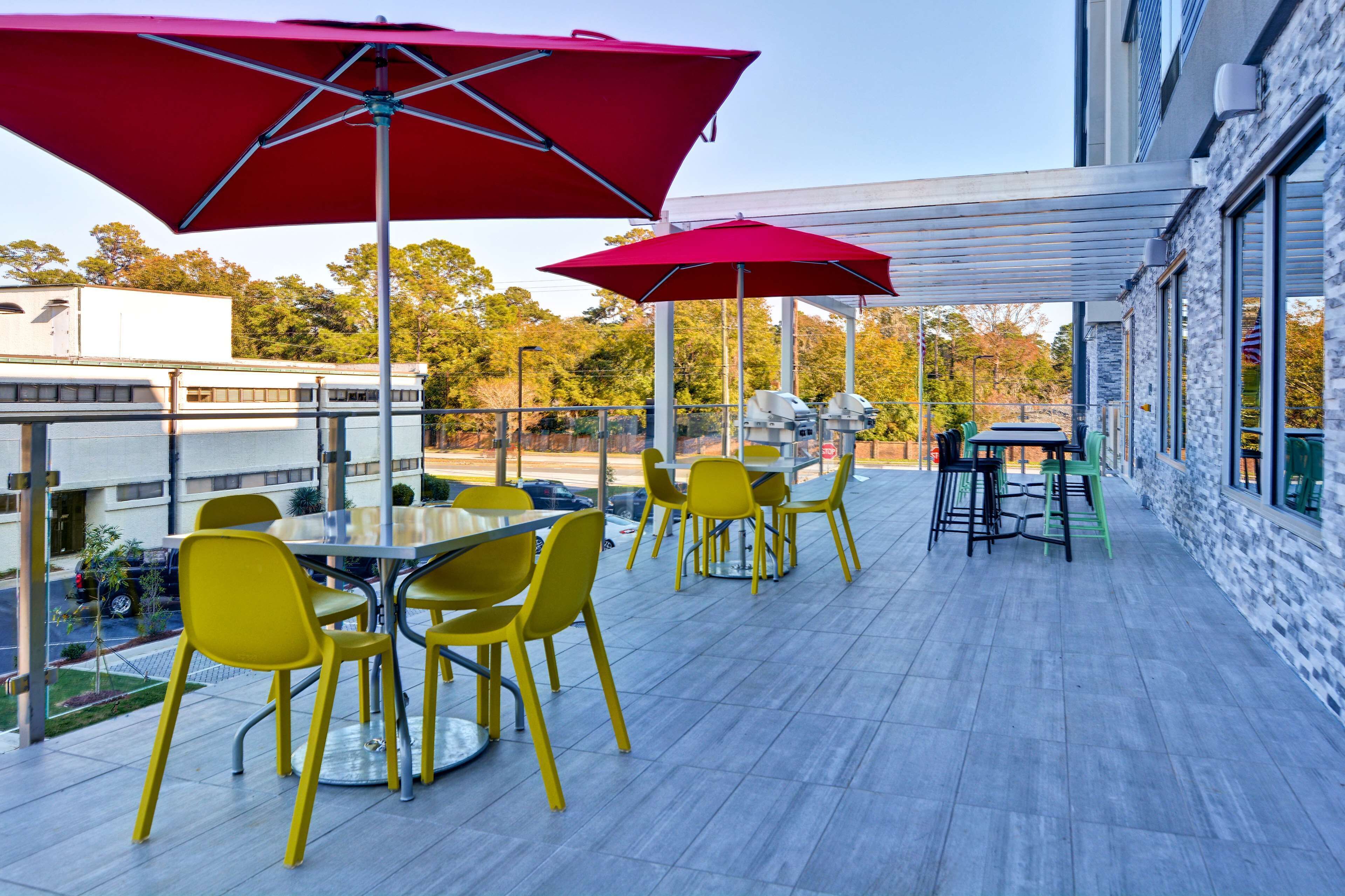 Eine Terrasse im Freien mit gelben Stühlen und roten Sonnenschirmen, einer Glasumlaufung und Blick auf Bäume sowie ein Gebäude im Hintergrund.