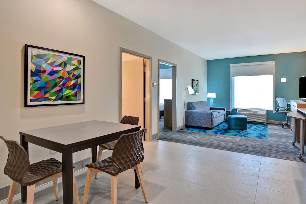 Une chambre d’hôtel moderne comprenant un coin repas, un espace de vie avec un canapé et un pouf, une grande fenêtre et un tableau abstrait aux couleurs vives.