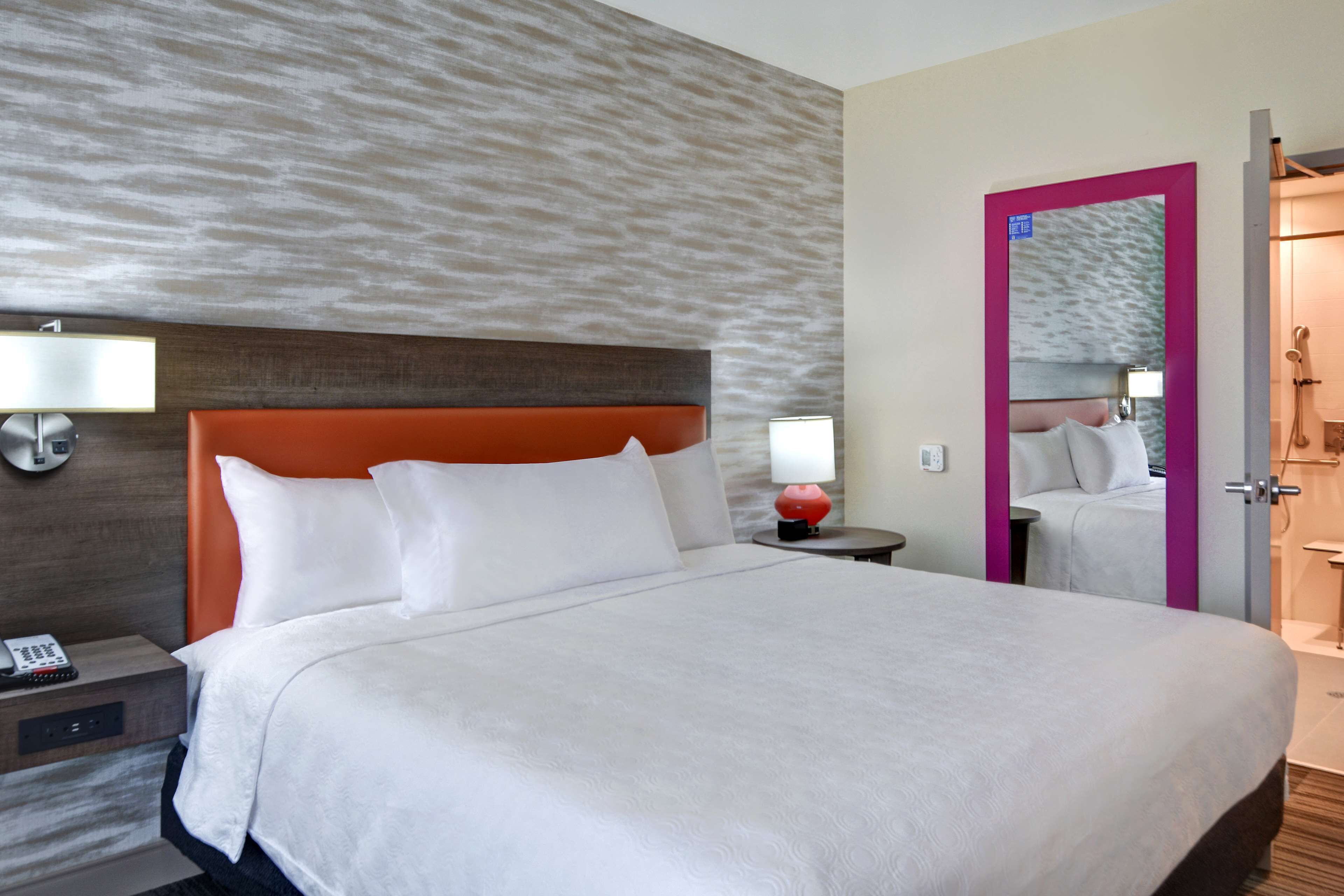A hotel room with a large bed, a nightstand with a lamp, and a bathroom visible through an open door.