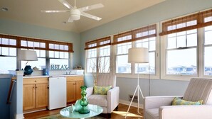 Amplia sala de estar con área de cocina, ventanas grandes con persianas, ventilador de techo y vista al vecindario.
