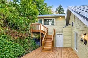 A two-story house with a wooden deck, a front door, and a staircase leading to a balcony. - Whidbey Island Beachfront Home | Hot Tub & Casita (Clinton)