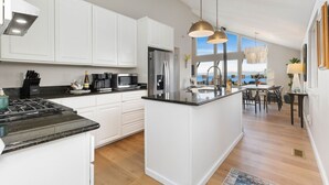 Una cocina moderna con armarios blancos, una isla central, electrodomésticos de acero inoxidable y vistas al exterior.