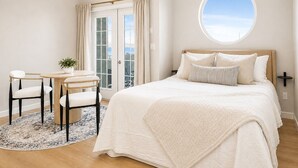 Un dormitorio con una cama grande, un ventanal redondo, una mesa de comedor con sillas y vistas al exterior a través de una puerta.
