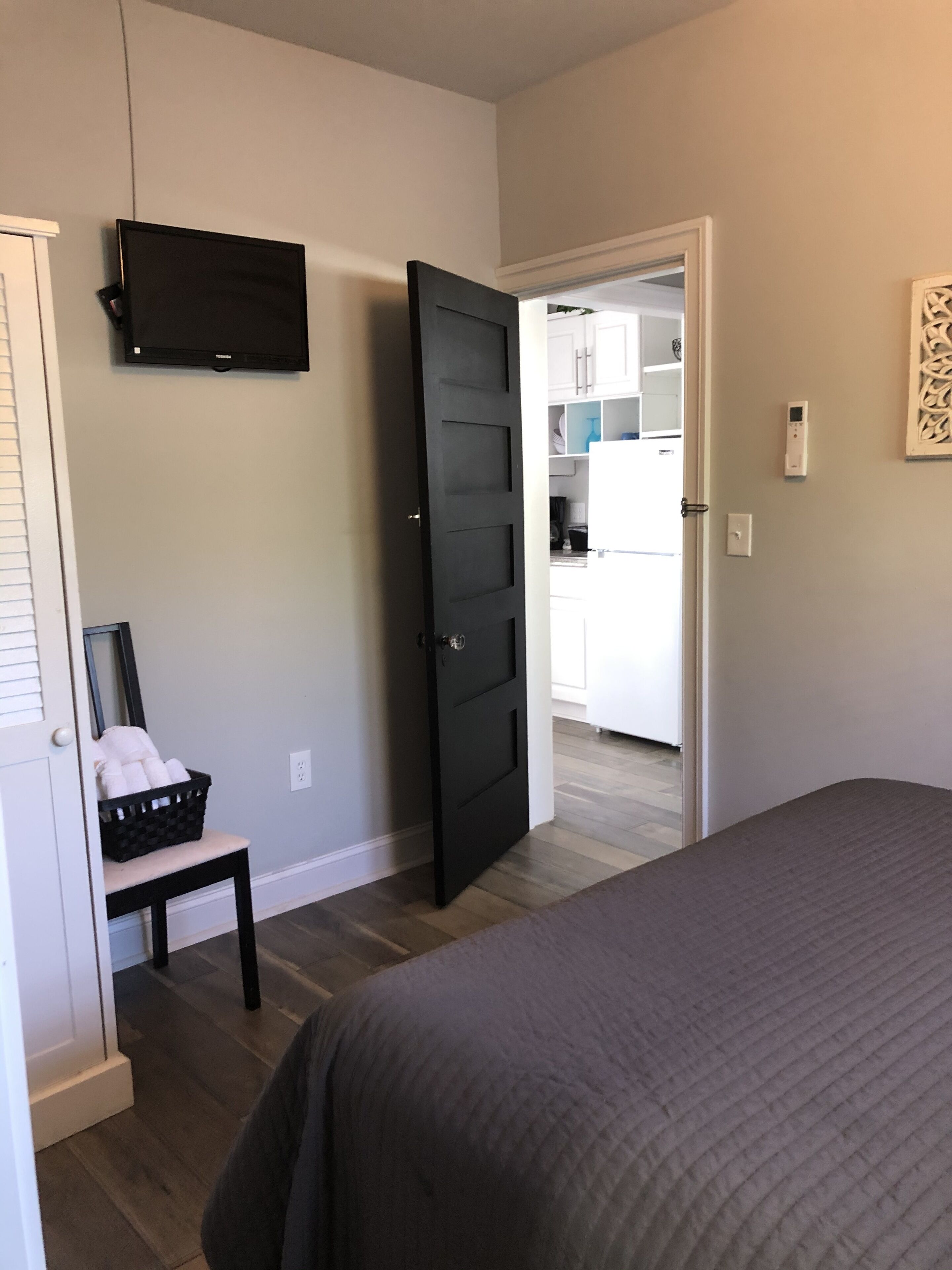 A bedroom with a bed, a television mounted on the wall, a door leading to a kitchen, and a small table with a basket.