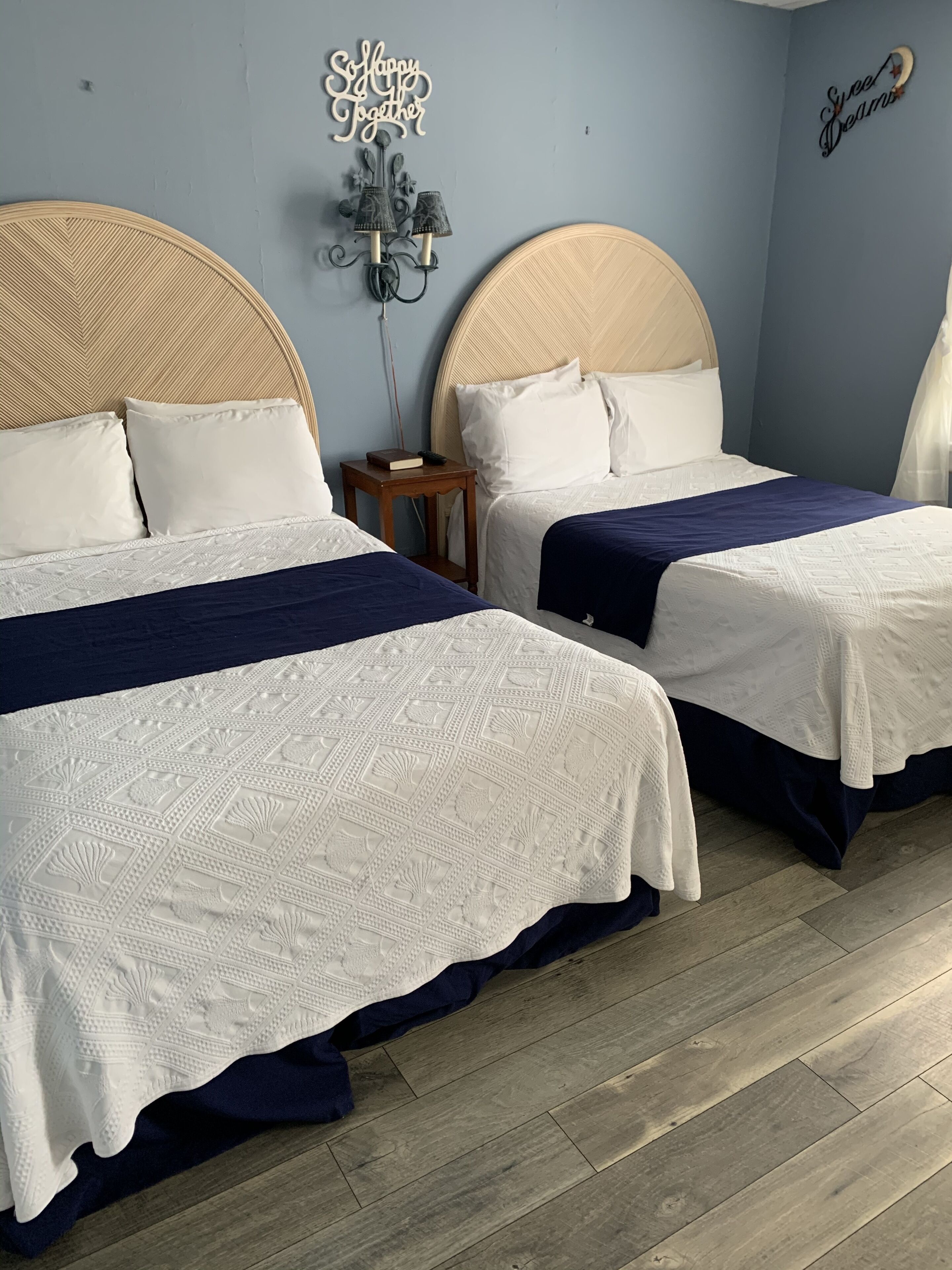 Hotellihuone, jossa on kaksi sänkyä, molemmissa pääty sekä pieni yöpöytä, jossa on kirja ja kaukosäädin, ja seinälle asennettu valaisin.