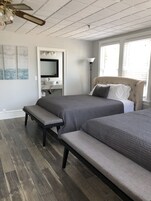 Un dormitorio con dos camas, un banco, un tocador con espejo, un ventilador de techo y vistas al exterior a través de la ventana.