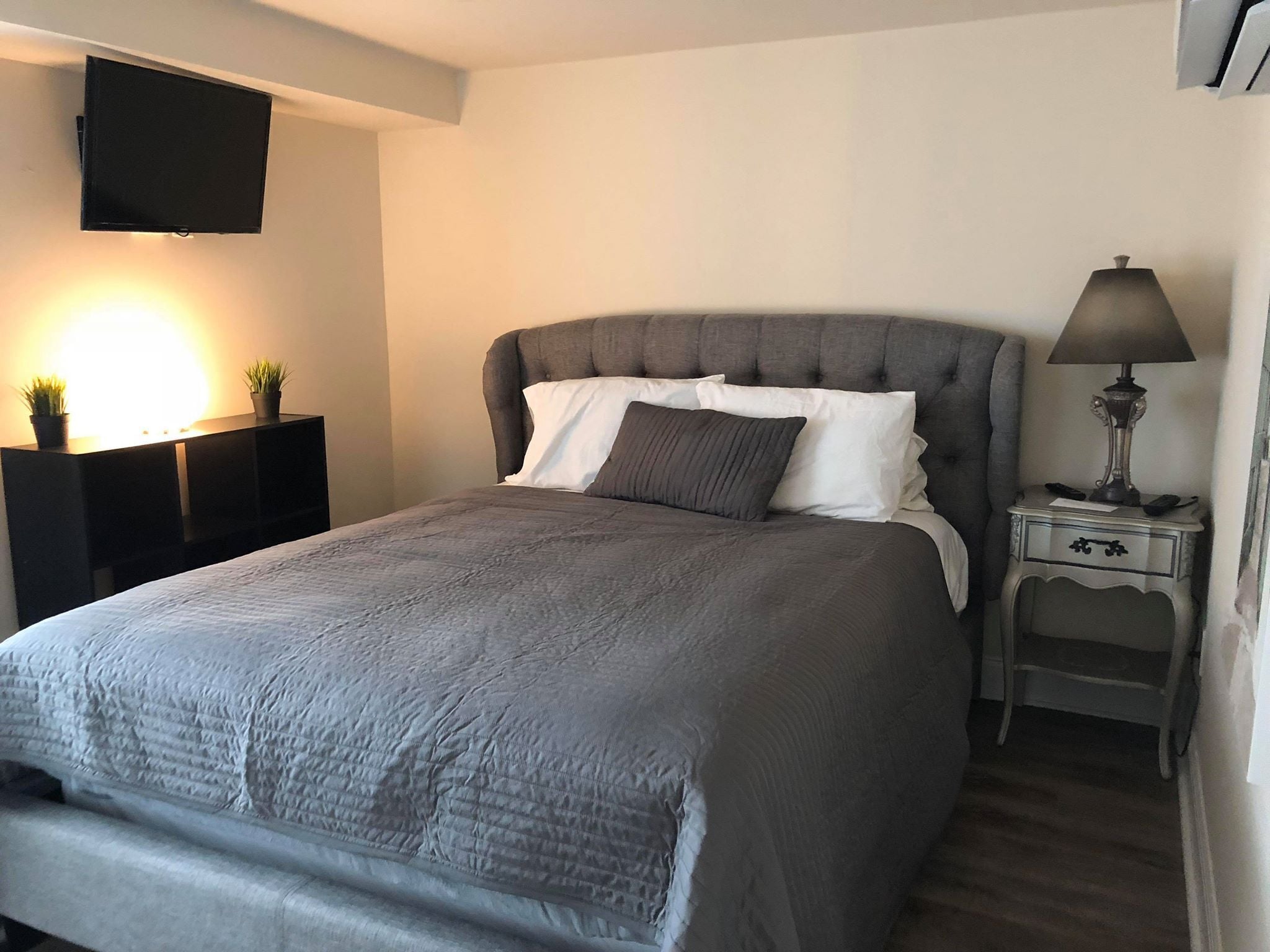 A neatly made bed with a grey quilt, a bedside lamp, and a flat-screen TV mounted on the wall.