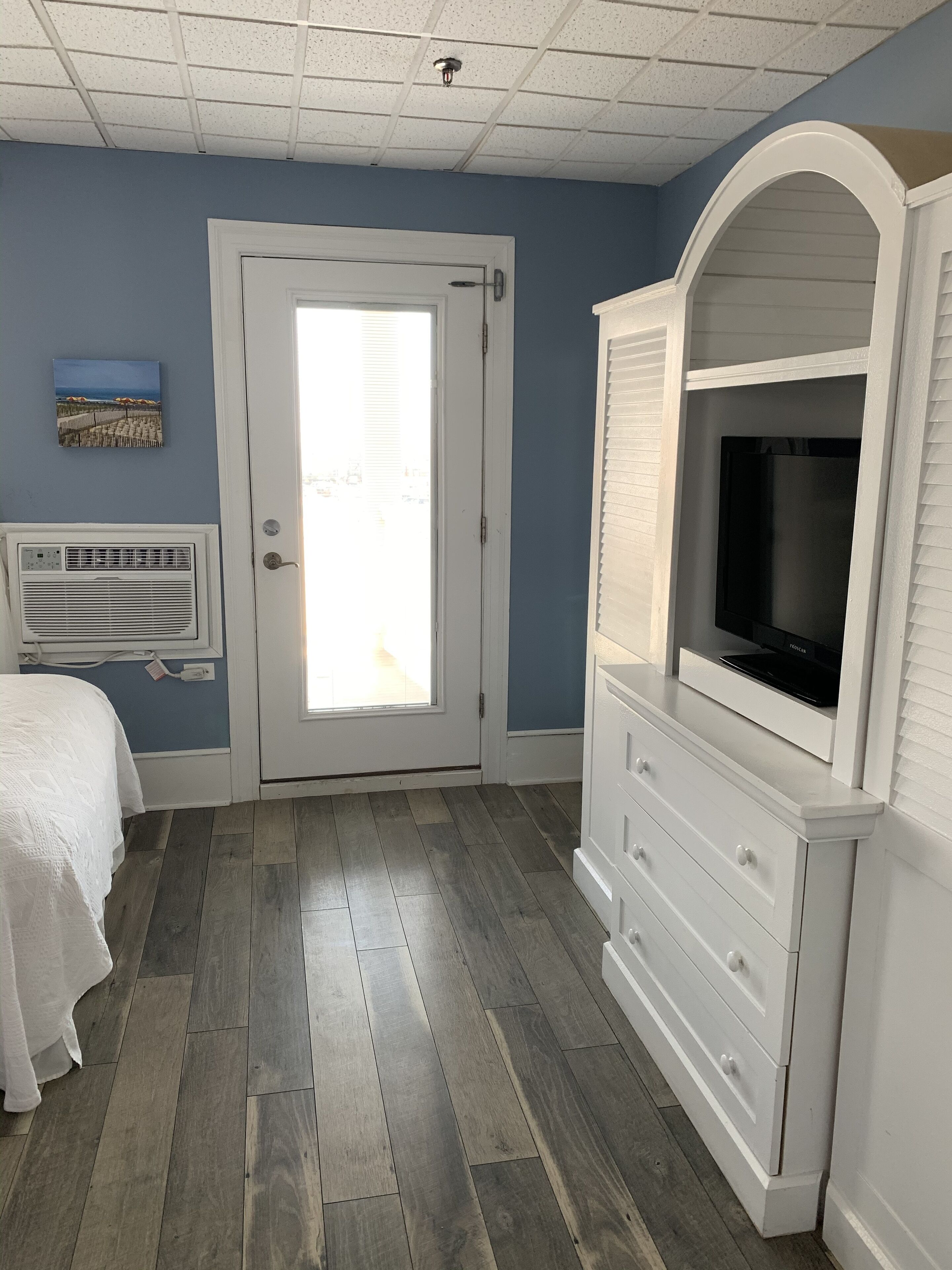 Camera da letto con porta che dà su un balcone, una cassettiera bianca con televisore e un letto con copriletto bianco.