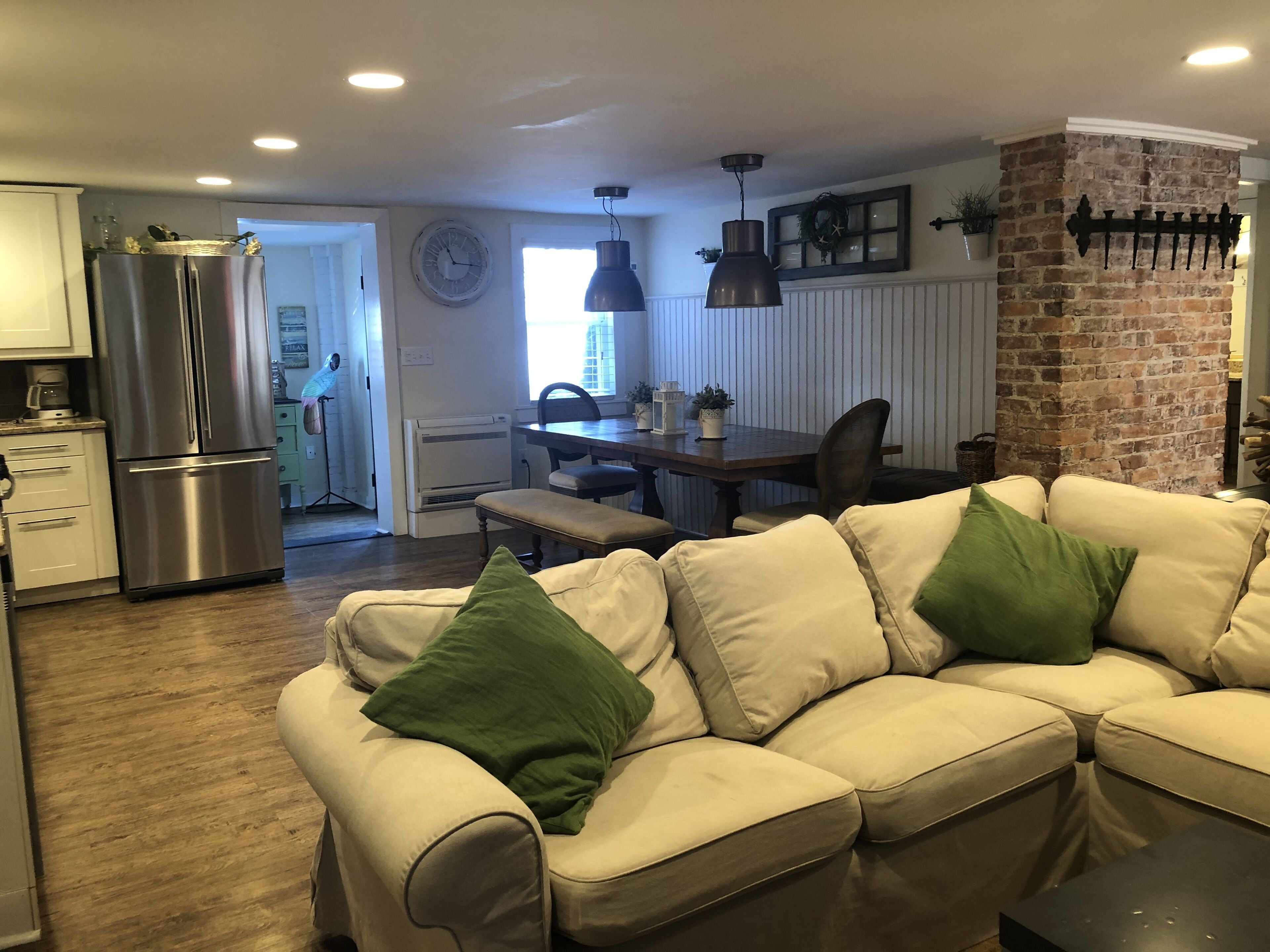 A modern kitchen with a stainless steel refrigerator, a dining area with a wooden table and chairs, and a living room with a beige sofa and green cushions.