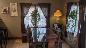 Un coin repas avec une table à plateau en verre, des chaises en bois, un ventilateur de plafond et une fenêtre agrémentée de rideaux à motifs floraux.