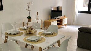 A dining area with a white table set for four, surrounded by white chairs. A television is mounted on the wall, and a sofa is visible in the background. - Fuengirola, 1 min. walk from the beach  (Fuengirola)