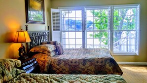 Une chambre avec une grande fenêtre, un lit recouvert d’une couvre-lit à motifs, une table de chevet avec une lampe et un tableau encadré au mur.