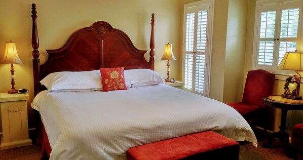 Una cama bien hecha con cabecera de madera, dos lámparas de noche, una silla roja y una mesita con lámpara.
