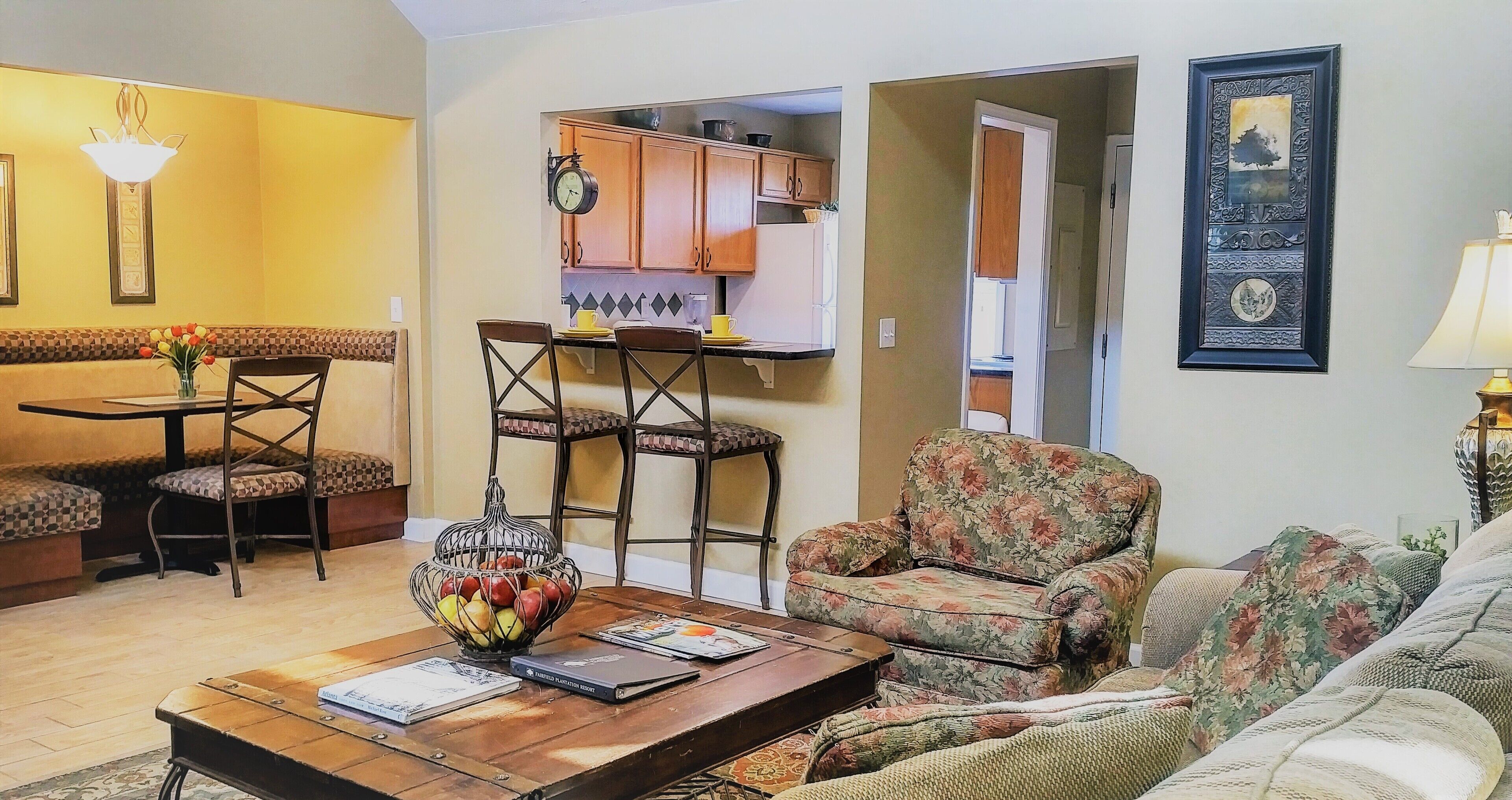 A living room with a sofa, chairs, a coffee table with books, and a kitchen area in the background.