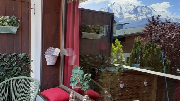 A balcony with a view of snow-capped mountains, a wicker chair, a red cushion, and a glass table with a candle. - Studio in the heart of Combloux village (Combloux)
