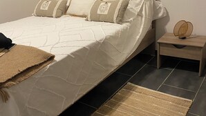 A bed with a white quilt, multiple pillows, a wooden nightstand, a woven rug, and a wall switch. - Ground floor apartment, T2 type, sea view. (Santa-Reparata-Di-Balagna)