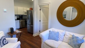 Ein Wohnzimmer mit einem weißen Sofa, einem Couchtisch und einem runden Spiegel an der Wand. Im Hintergrund ist eine Küche mit weißen Schränken und einem Edelstahl-Kühlschrank erkennbar.
