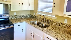 A compact kitchen with white cabinets, a granite countertop, stainless steel appliances, and a window with blinds. - Lakeview Cottage Near Boat Ramp (Burnet)