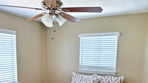 A bedroom with a patterned bedspread, a ceiling fan, and two windows with blinds. - Lakeview Cottage Near Boat Ramp (Burnet)