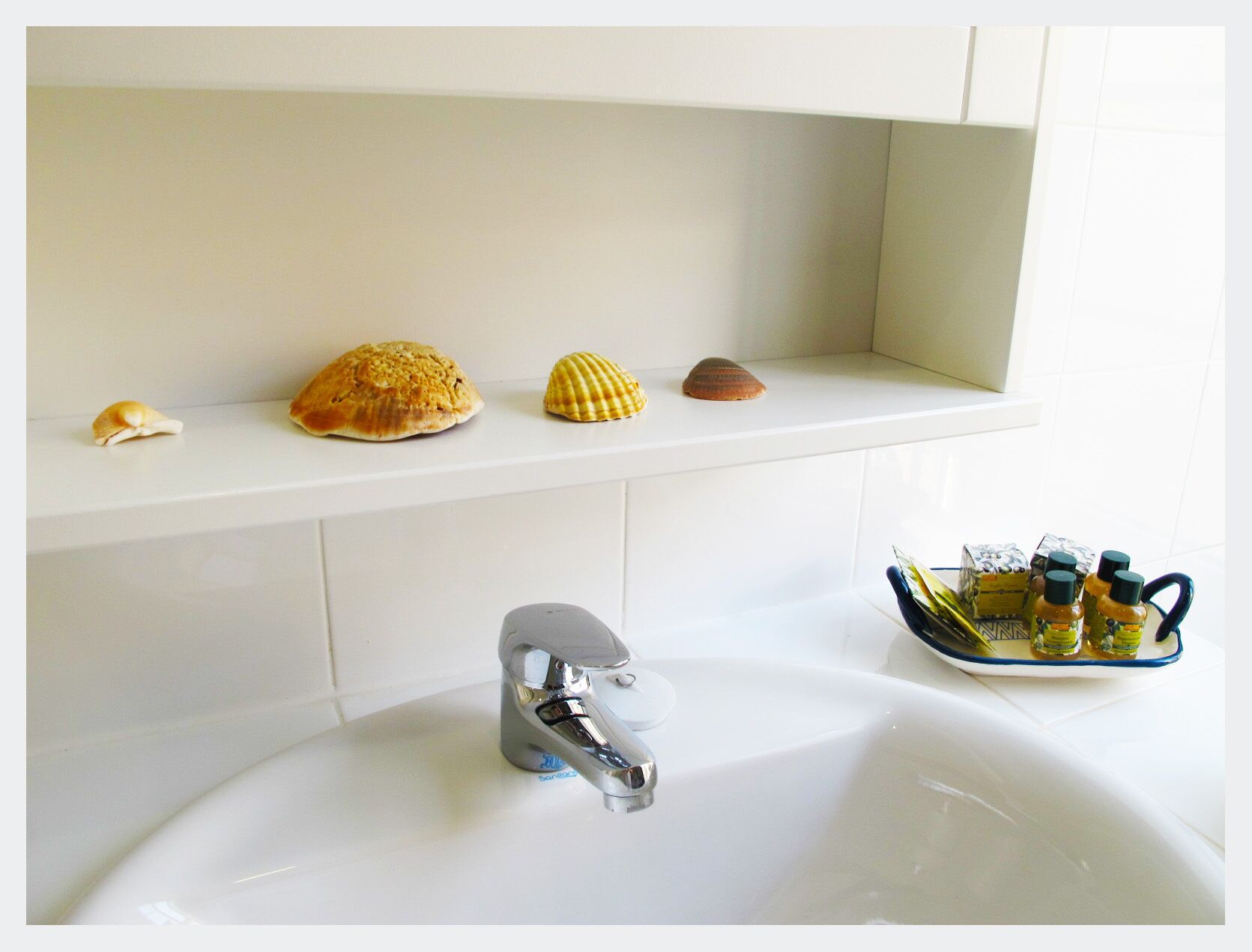Une salle de bain avec un lavabo blanc, un robinet chromé et une étagère au-dessus du lavabo où sont exposés divers coquillages et articles de toilette.