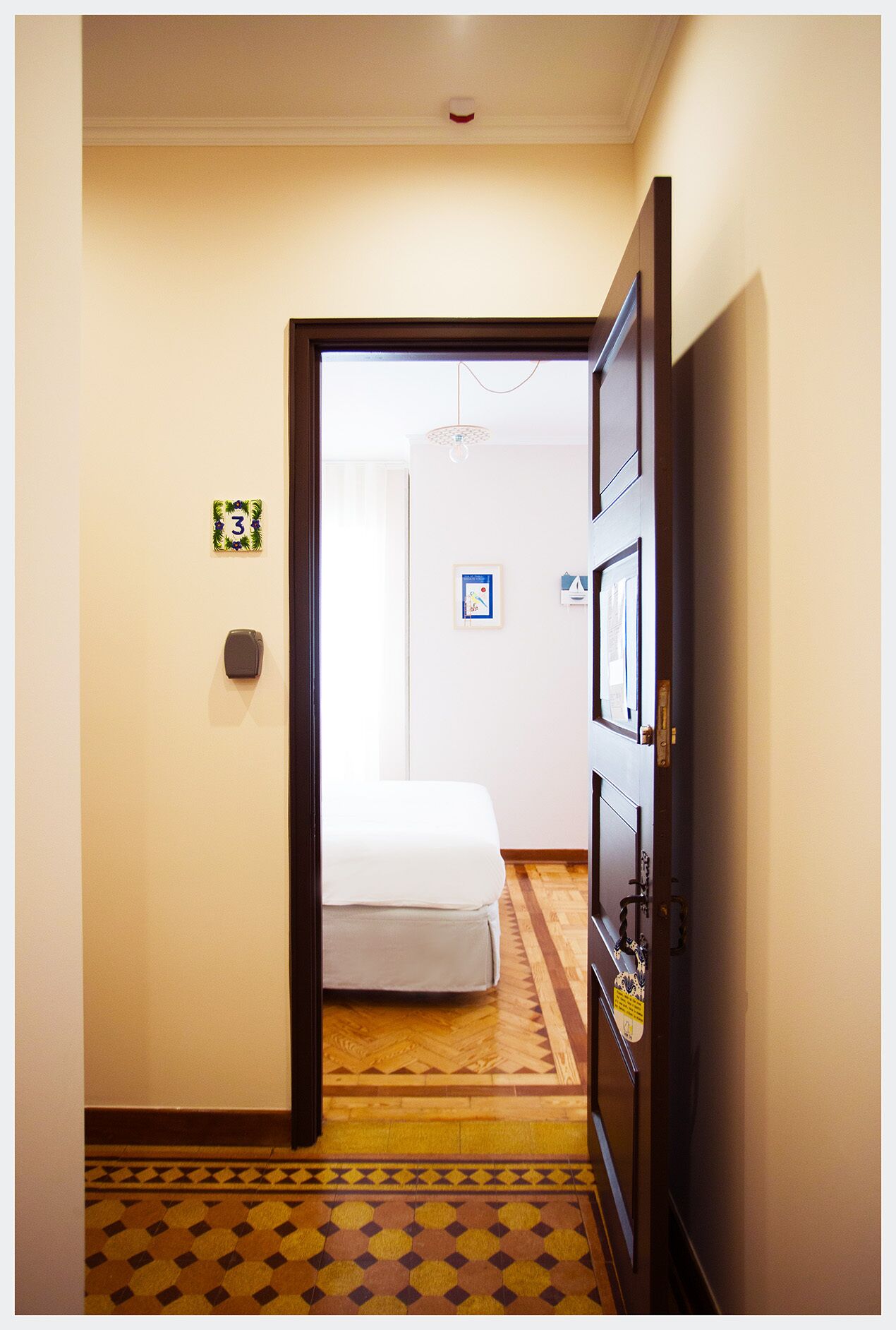 A narrow hallway with a patterned floor, a white door frame, a white door, and a bed in the background.