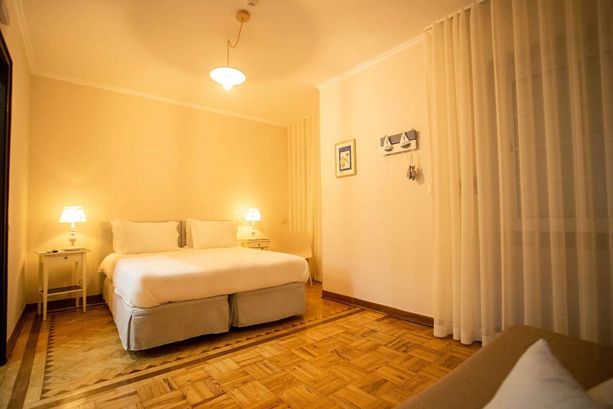 A hotel room with a bed, two bedside lamps, a wooden floor, and a window with curtains.