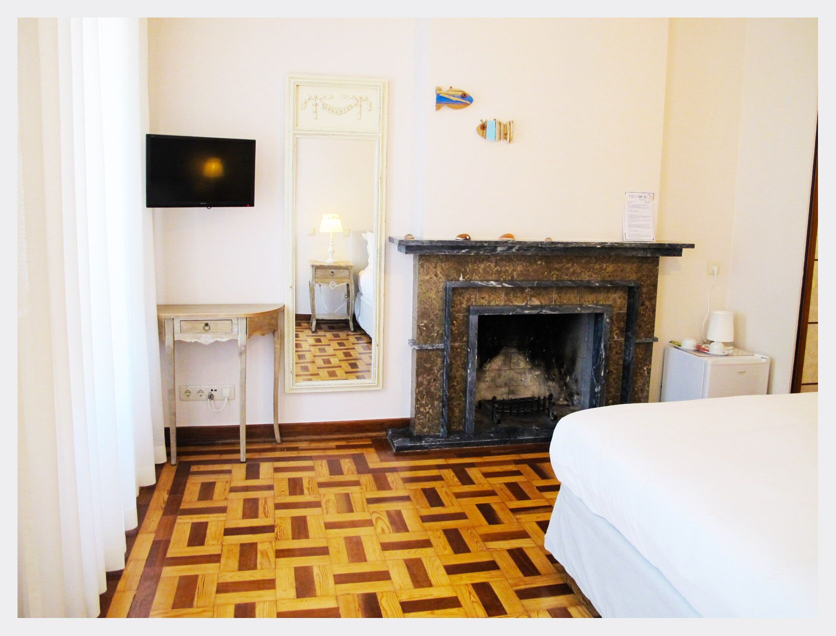 A hotel room with a wooden parquet floor, a bed, a small bedside table, a mirror, a fireplace, and a television mounted on the wall.