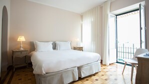 A hotel room with a large bed, two bedside tables with lamps, a wooden floor, and a balcony with a chair.
