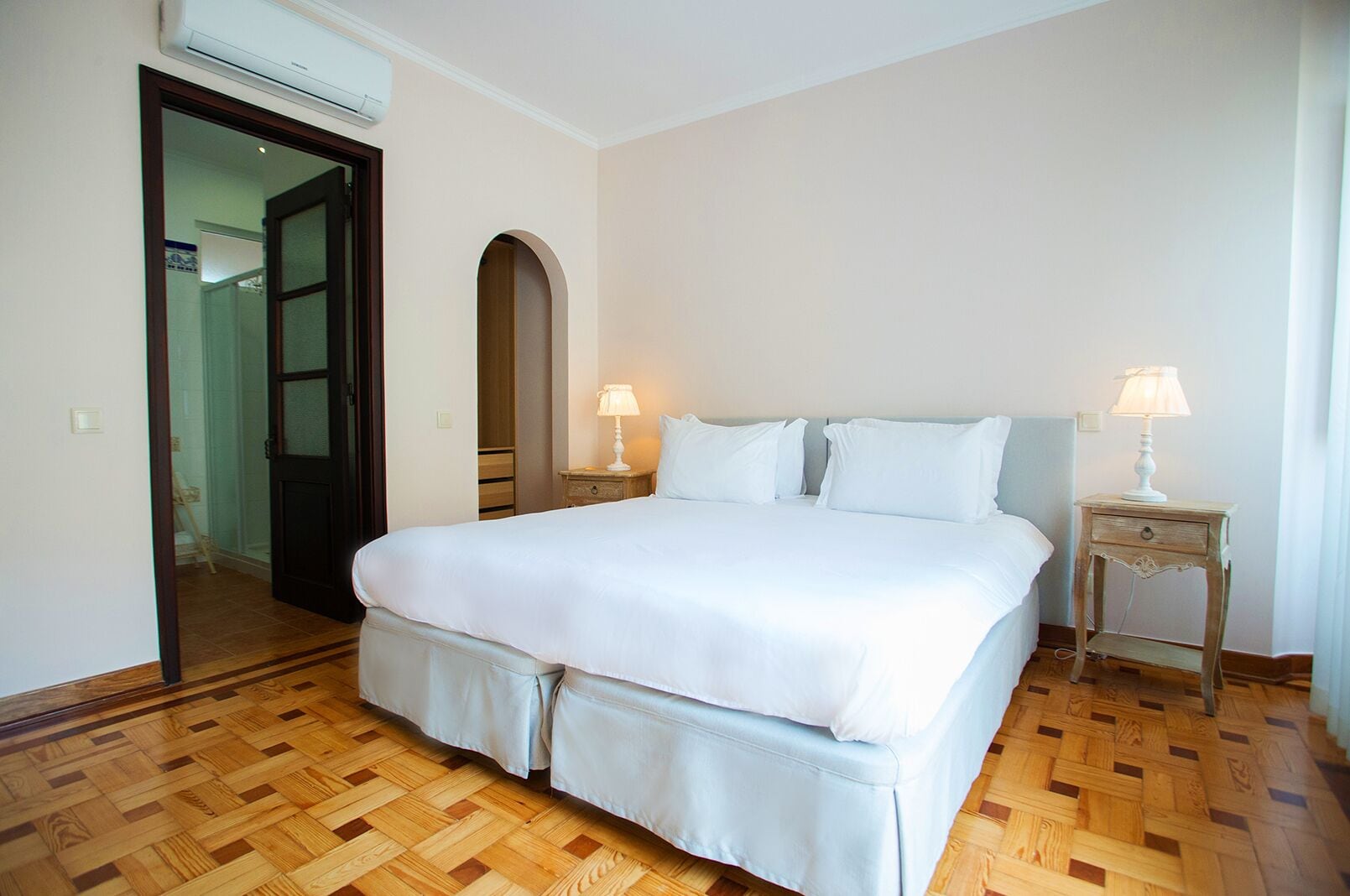 A bedroom with a large bed, two bedside tables with lamps, a wooden floor, and a door leading to another room.