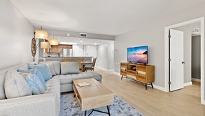 A modern living room with a sectional sofa, a coffee table, and a TV. There is a kitchen in the background with a dining area.