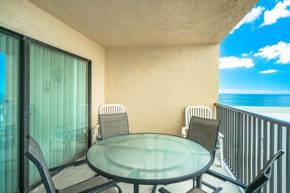 A balcony with a glass table and chairs, overlooking a beach.